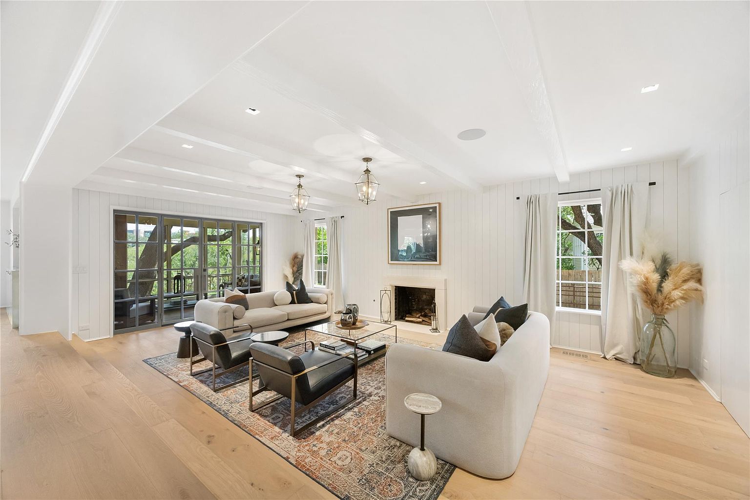 This is a bright and airy living room featuring white walls, exposed ceiling beams, and hardwood floors. The room is furnished with two sofas, two armchairs, a glass coffee table, and a decorative rug. Large windows and glass doors provide ample natural light and views of the outdoors, creating a welcoming and sophisticated atmosphere.