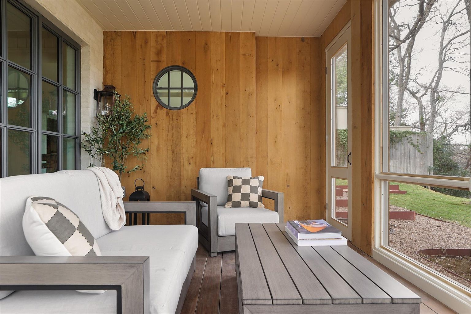 This image showcases a well-designed outdoor patio area, featuring comfortable seating arrangements with a light gray sofa and armchair, complemented by a wooden coffee table. The space is enclosed with wood paneling and large windows offering a view of the yard, creating a cozy and inviting atmosphere. A round window adds a unique architectural detail.