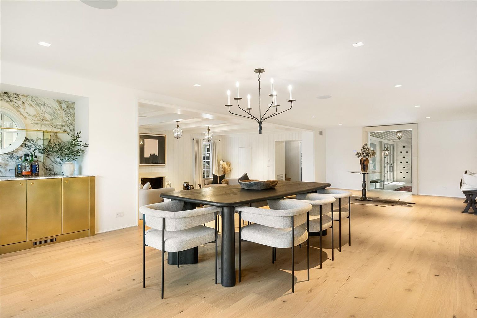 This is a bright and spacious dining room featuring a large, dark wood dining table with modern chairs. A stylish chandelier hangs above the table, providing ample lighting. The room has light wood floors and white walls, creating a clean and airy atmosphere, with a gold cabinet and marble backsplash adding a touch of luxury.