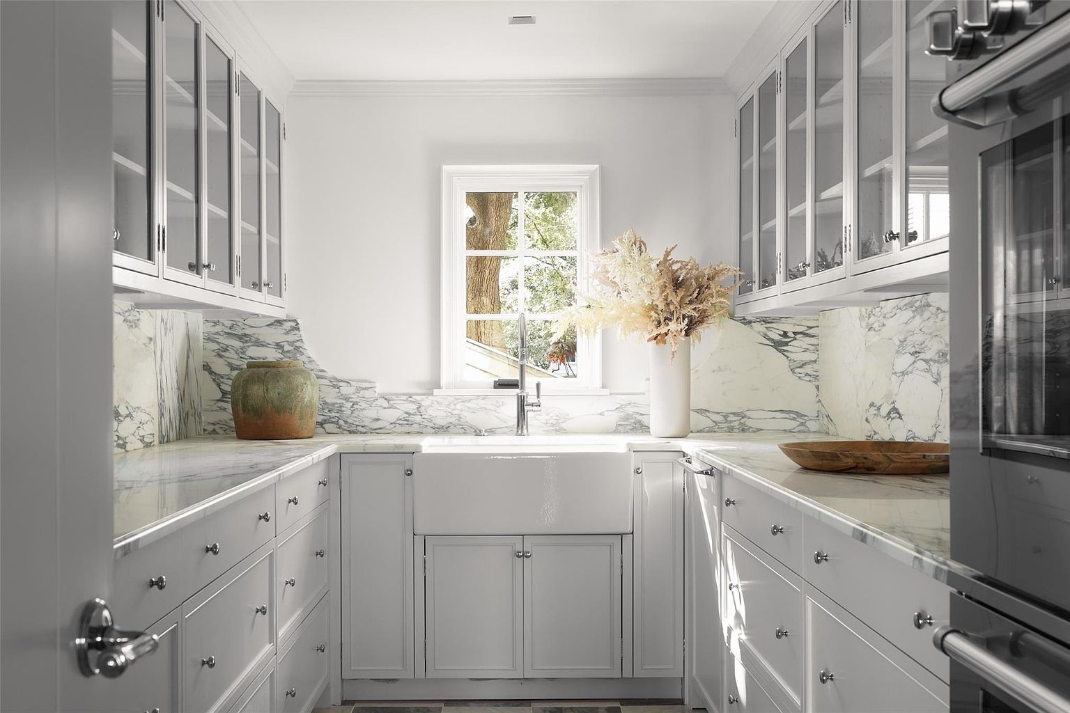 This is a bright and elegant kitchen featuring white cabinetry with glass fronts, marble countertops and backsplash, and a farmhouse sink centered under a window. The design incorporates classic elements with a clean, modern aesthetic, creating a sophisticated and functional space. Stainless steel appliances are visible, adding to the kitchen's high-end appeal.
