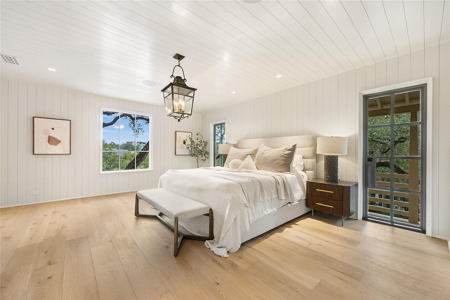 This is a bright and airy primary bedroom featuring a neutral color palette. The room has light wood flooring, white paneled walls, and a matching paneled ceiling. A large bed with a padded headboard is the focal point, complemented by a bench at the foot of the bed and a dark wood nightstand with a lamp. A lantern-style light fixture hangs from the ceiling, adding a touch of elegance.