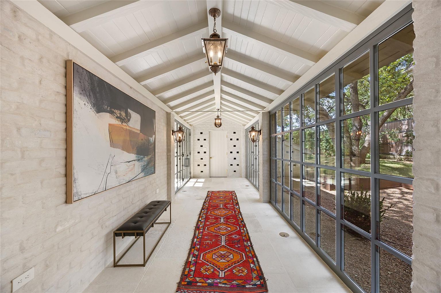This is an interior shot of a long hallway featuring white brick walls, a white vaulted ceiling with exposed beams, and a light-colored tile floor. A large abstract painting hangs on the left wall above a bench, while a series of windows line the right wall, offering views of the exterior. An orange and red patterned rug runs down the center of the hallway, leading to a white door at the end.