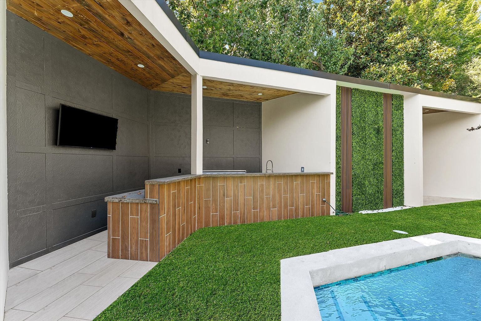 This image showcases a modern outdoor kitchen and pool area. The kitchen features a wooden bar, a built-in grill, and a mounted television, all under a stylish wooden-paneled roof. The pool is surrounded by lush green grass, creating a luxurious and inviting backyard space perfect for entertaining.