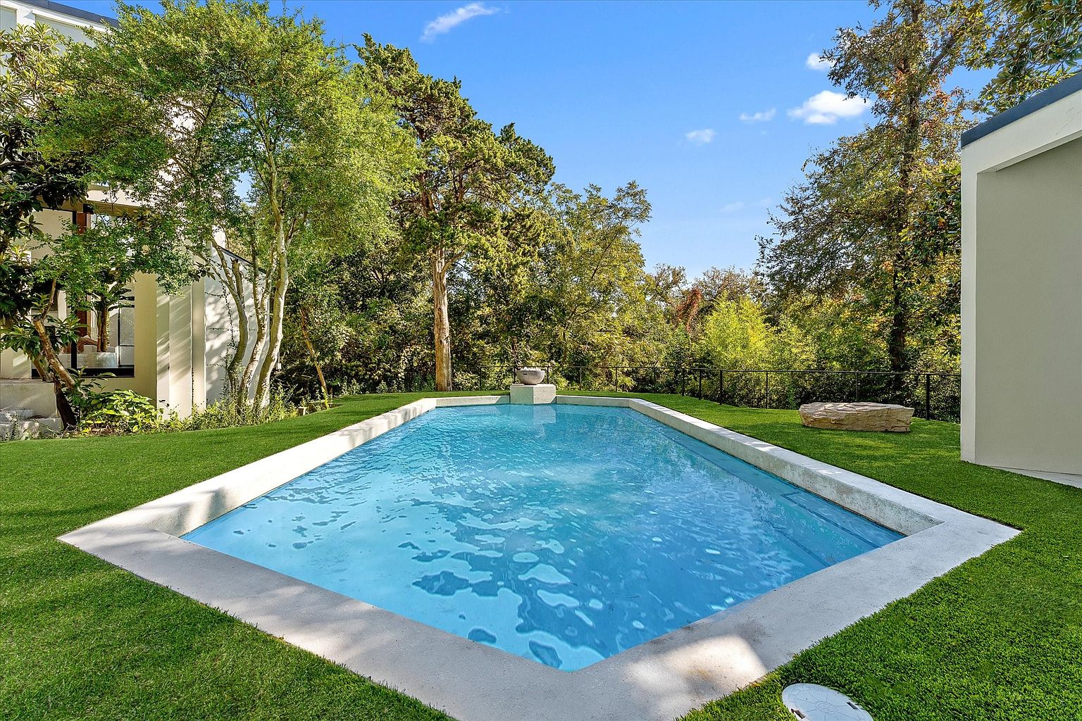 This image showcases a rectangular swimming pool with a modern, minimalist design, surrounded by lush green grass. The pool's clear blue water reflects the sky, and the surrounding trees create a serene and private backyard oasis. A contemporary building is visible on the right, adding to the property's upscale appeal.