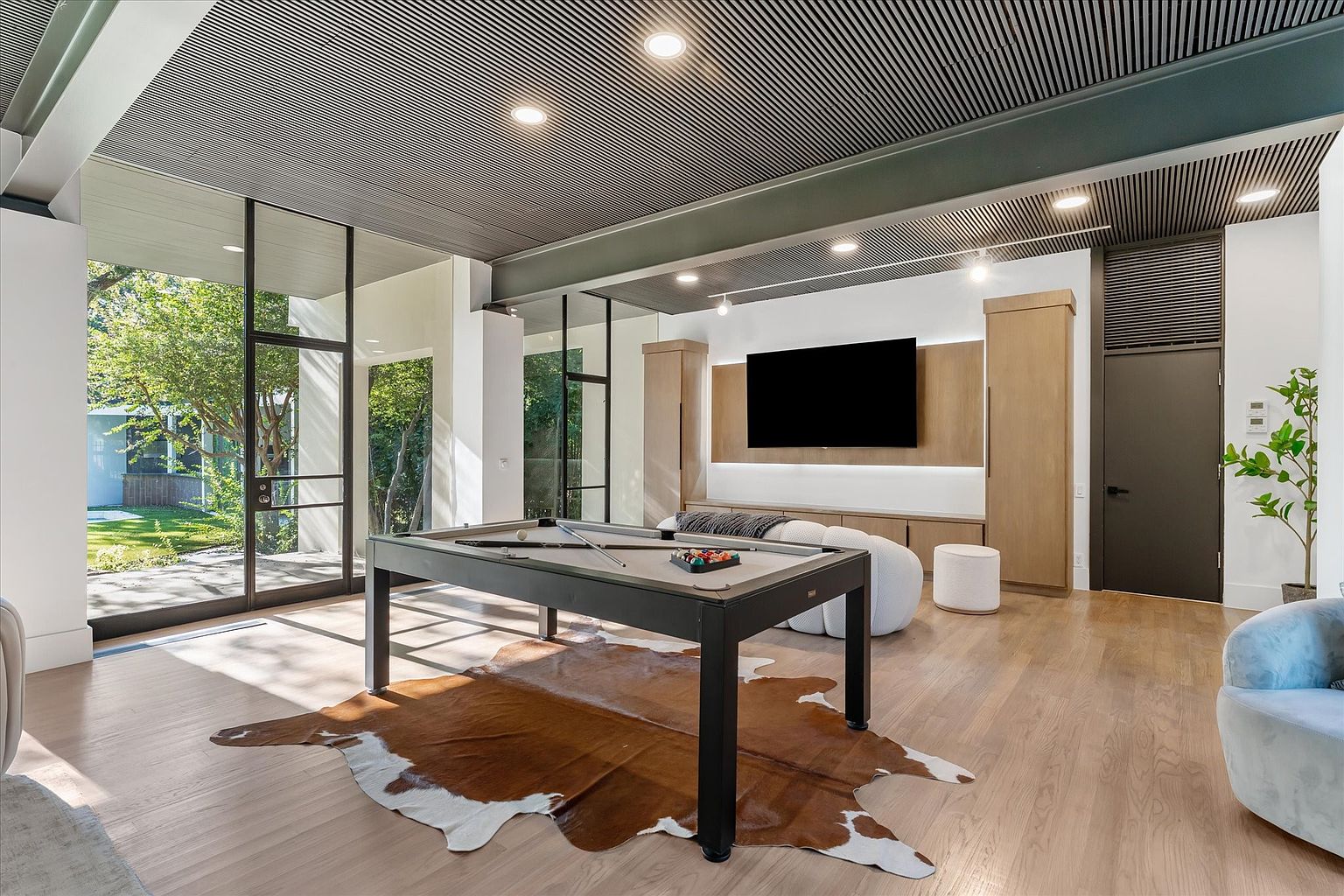 This interior shot showcases a modern living room with a unique ceiling design and large windows providing ample natural light. A pool table sits atop a cowhide rug, adding a touch of rustic charm to the contemporary space. The room also features a large television, built-in cabinetry, and stylish seating, creating a comfortable and visually appealing entertainment area.