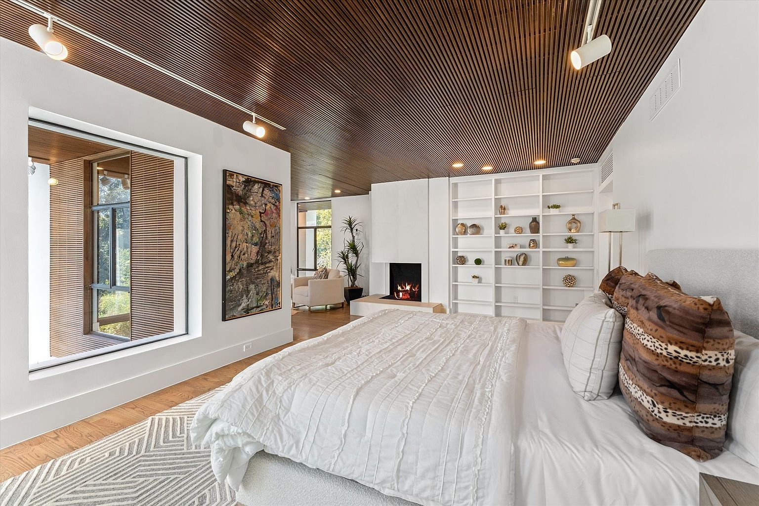 This is a primary bedroom featuring a modern design with a unique wooden slat ceiling and track lighting. The room includes a large bed with white linens and decorative pillows, a fireplace with a white surround, and a built-in bookshelf. A large window offers natural light, and an abstract painting adds a touch of color to the white walls.