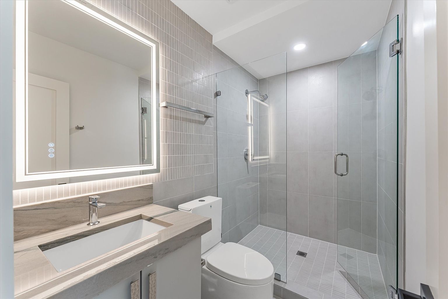 This is a modern bathroom featuring a floating vanity with a light countertop and a rectangular sink. A large, illuminated mirror hangs above the vanity, and a toilet is positioned next to it. The shower is enclosed with glass panels and features gray tiling, creating a sleek and contemporary look.