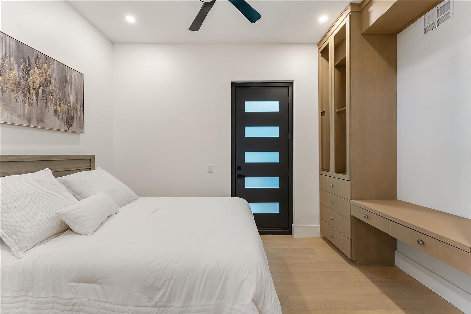 This is a well-lit primary bedroom featuring a king-size bed with white linens, a modern black door with frosted glass panels, and a custom built-in wardrobe with a desk area. The room has light wood flooring and a neutral color palette, creating a serene and inviting atmosphere. An abstract painting hangs above the bed, adding a touch of artistic flair.