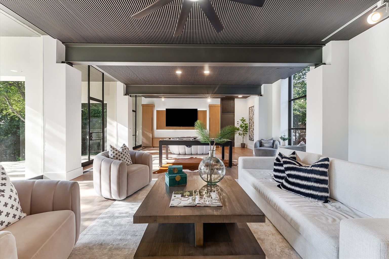 This is an interior shot of a modern living room. The room features a dark, slatted ceiling with recessed lighting, white walls, and large windows that provide natural light. The furniture includes a light-colored sofa with striped pillows, two armchairs, and a large wooden coffee table with decorative items. A game table is visible in the background.