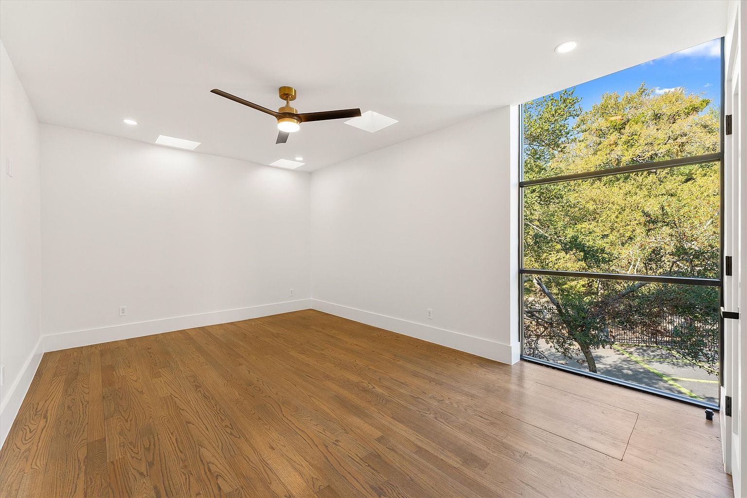 This is an interior shot of a modern bedroom featuring hardwood floors, white walls, and a large window offering a view of lush greenery. The room is illuminated by recessed lighting and a ceiling fan, creating a bright and airy atmosphere. The minimalist design and natural light enhance the room's appeal, making it feel spacious and inviting.