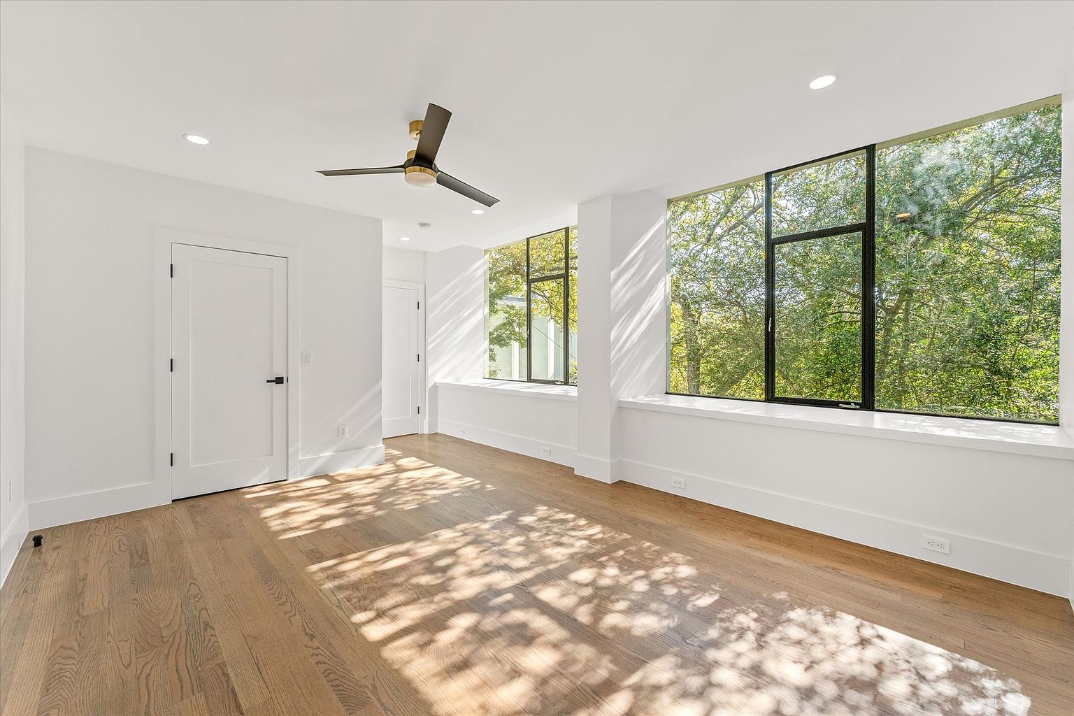This is a bright and airy primary bedroom featuring hardwood floors and large windows that provide ample natural light and views of the surrounding trees. The room is painted in a neutral color, enhancing the sense of spaciousness. A modern ceiling fan adds a touch of contemporary style.