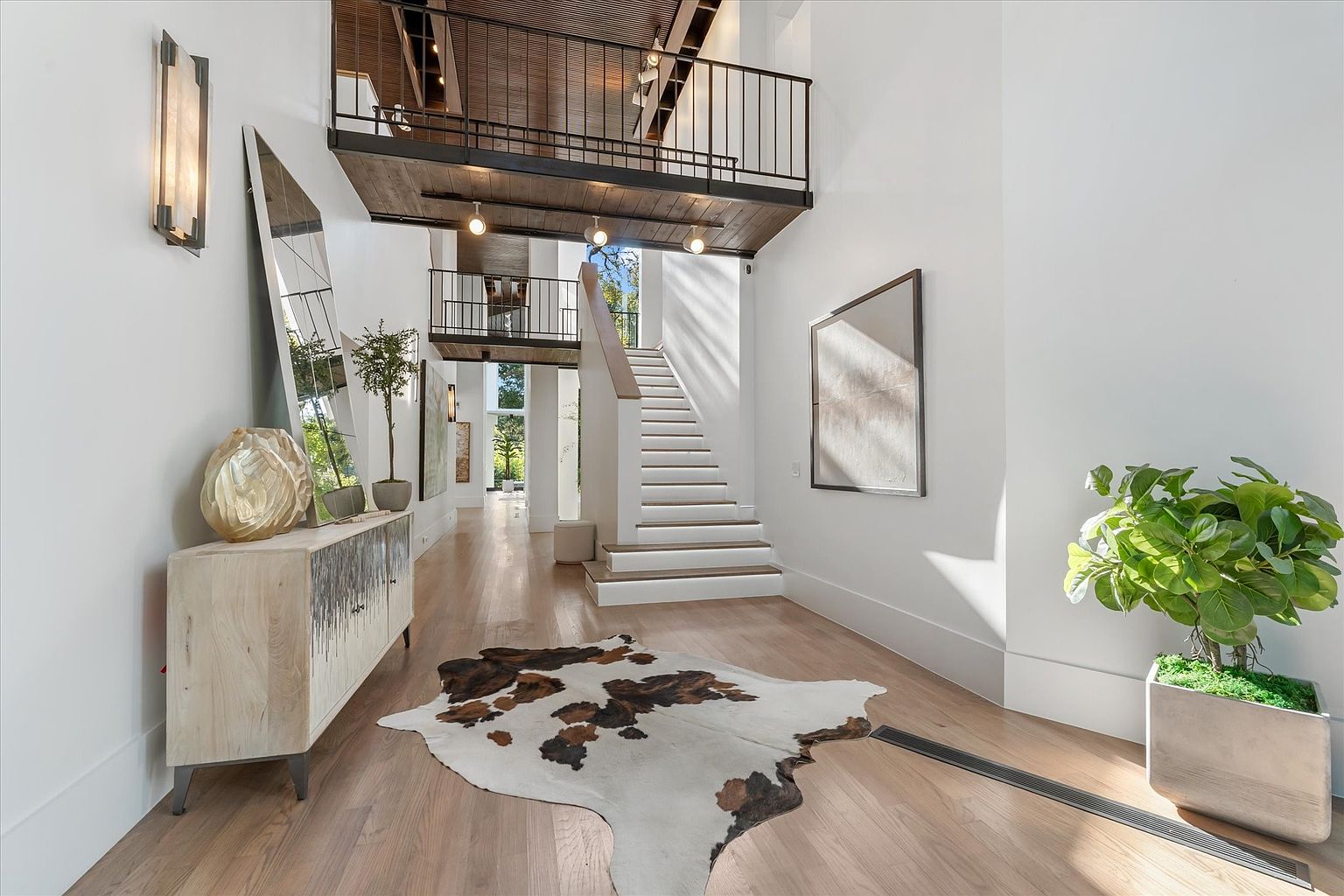 This is an interior shot of a grand hallway featuring a staircase leading to a second-story balcony. The hallway is decorated with modern art, a cowhide rug, and a stylish console table with decorative objects. The space is bright and airy, with natural light streaming in, creating a welcoming and luxurious atmosphere.