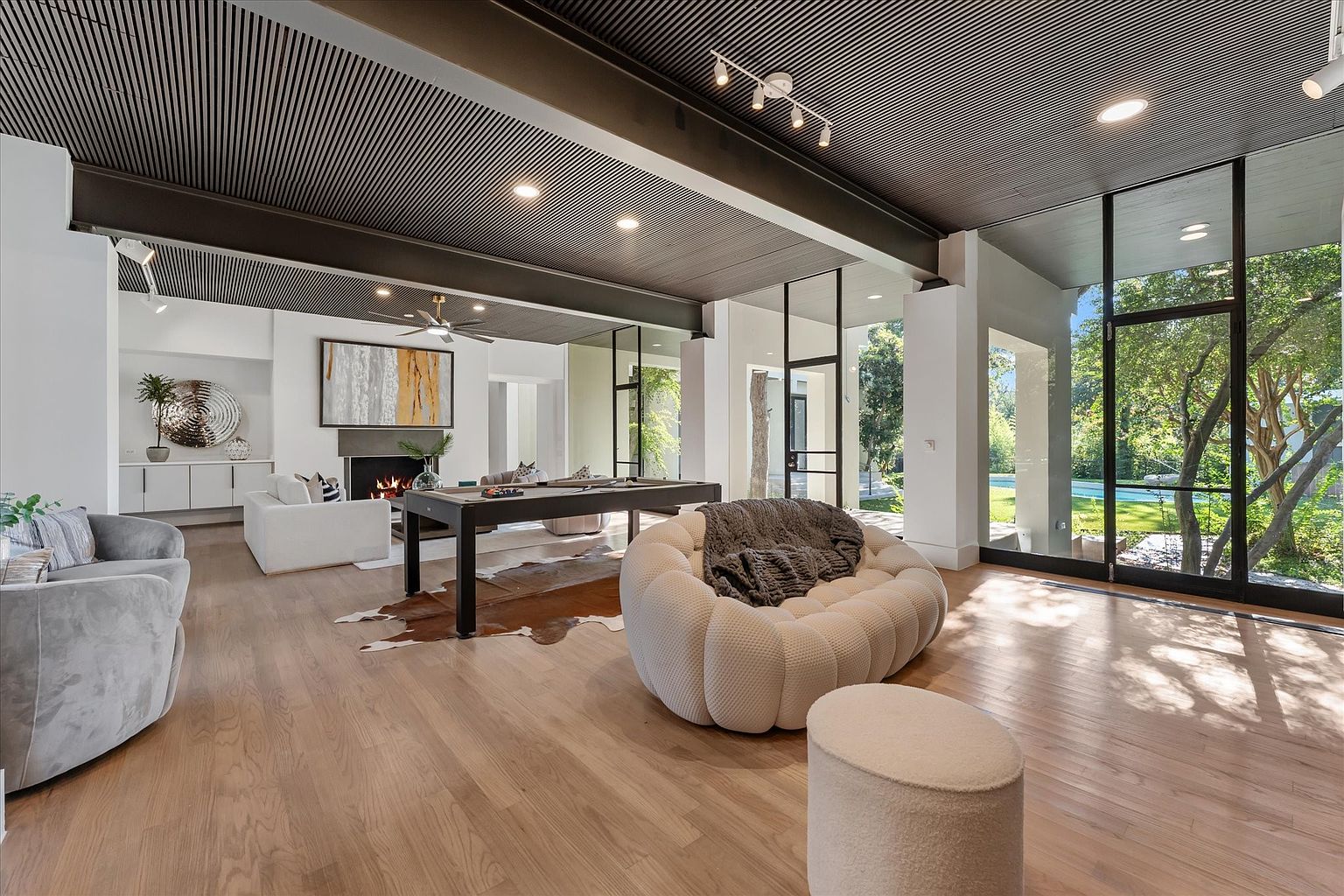 This is a spacious and modern living room featuring hardwood floors, a unique slatted ceiling, and large windows offering ample natural light and views of the outdoors. The room includes a fireplace, a pool table, and comfortable seating arrangements, creating a versatile space for relaxation and entertainment. The design incorporates a blend of contemporary and minimalist elements, enhancing the room's overall appeal.