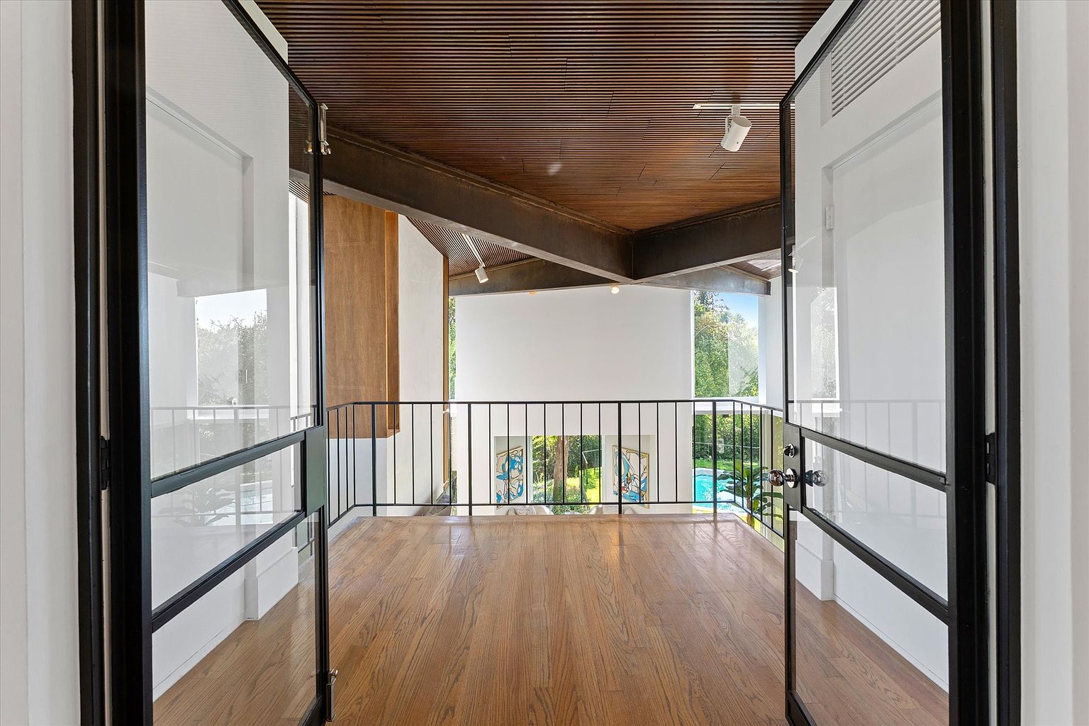This interior shot showcases a hallway or landing area with hardwood floors and a black metal railing overlooking a lower level. Large glass doors with black frames open to the space, providing ample natural light and a view of the exterior. The ceiling features a unique wood slat design, adding architectural interest.