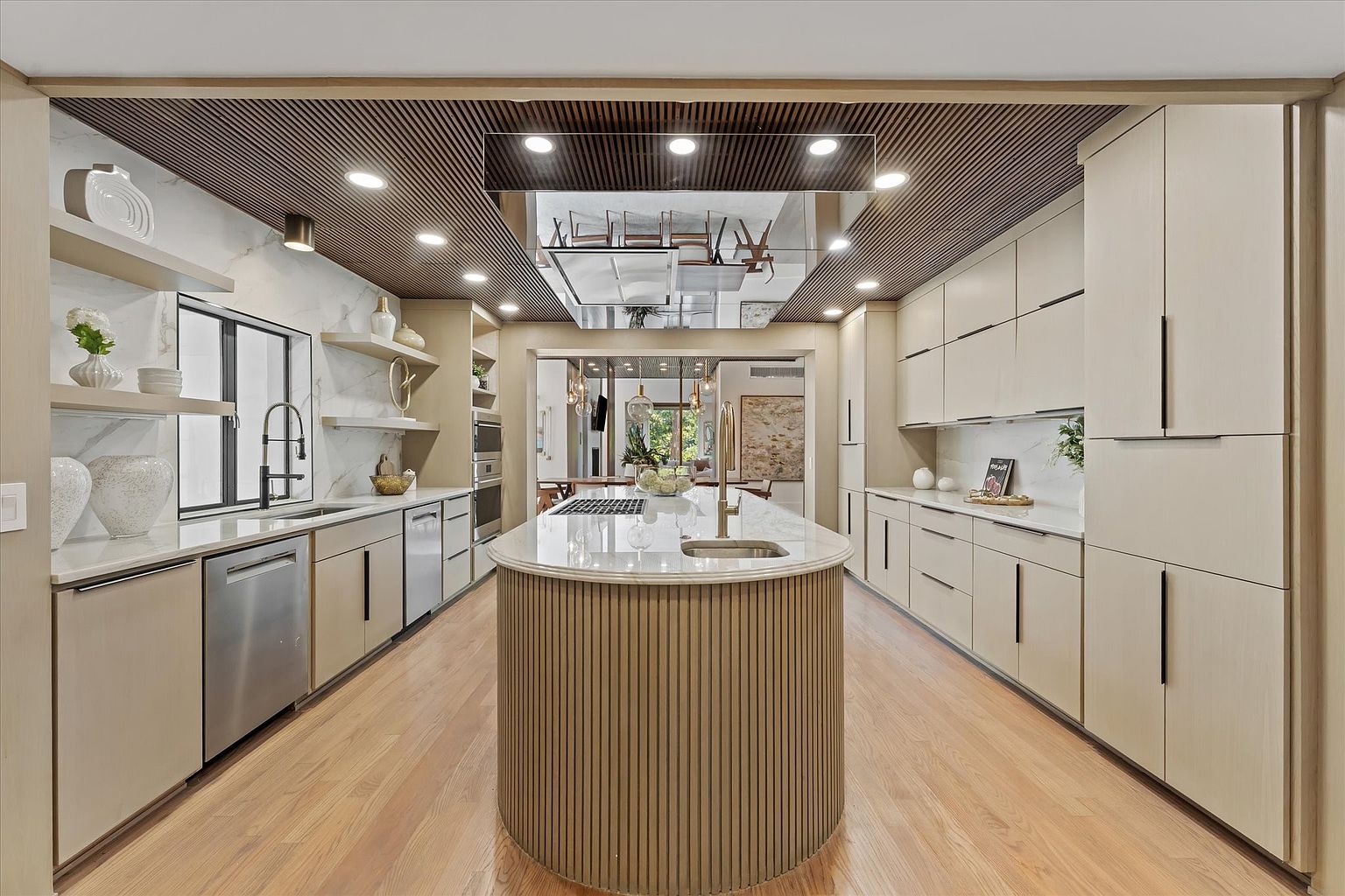This is a wide shot of a modern kitchen featuring light wood cabinetry, stainless steel appliances, and a unique island with a rounded design and light wood paneling. The ceiling has a striking wood slat design with recessed lighting and a mirrored section above the island. The hardwood floors add warmth to the space, and the open doorway leads to another room, creating a sense of flow.