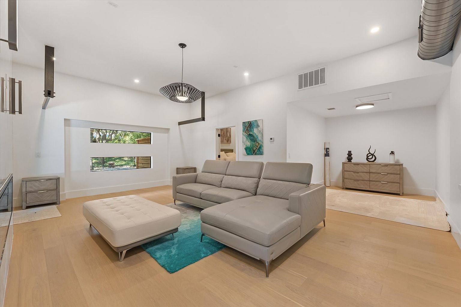 This is an interior shot of a modern living room featuring a gray leather sectional sofa and a matching ottoman on a light wood floor. A teal rug adds a pop of color, and a unique chandelier hangs from the ceiling. The room is bright and airy, with natural light coming through the windows.