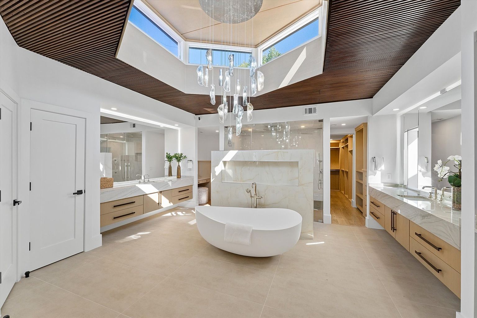 This is a luxurious primary bathroom featuring a freestanding white bathtub as a focal point, complemented by a modern chandelier and a skylight that floods the space with natural light. The bathroom includes dual vanities with light wood cabinetry and marble countertops, a glass-enclosed shower, and a walk-in closet, creating a spa-like atmosphere. The perspective is a wide shot to capture the spaciousness and high-end finishes.