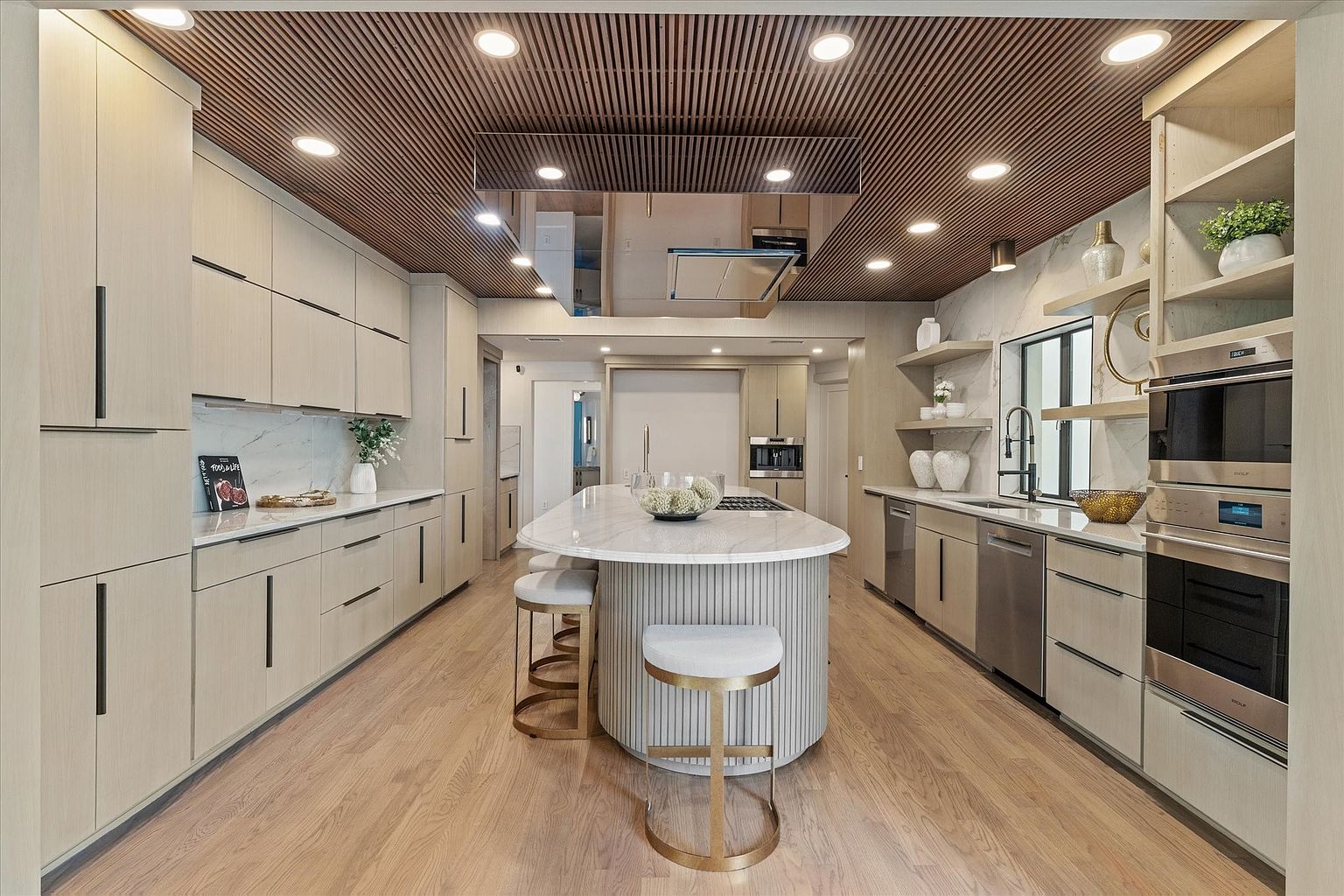 This is a bright and modern kitchen featuring light wood cabinetry with sleek, dark hardware and a large island with a marble countertop and seating. Stainless steel appliances and a unique wood-slatted ceiling add to the contemporary design. The kitchen is spacious and well-lit, creating an inviting atmosphere.