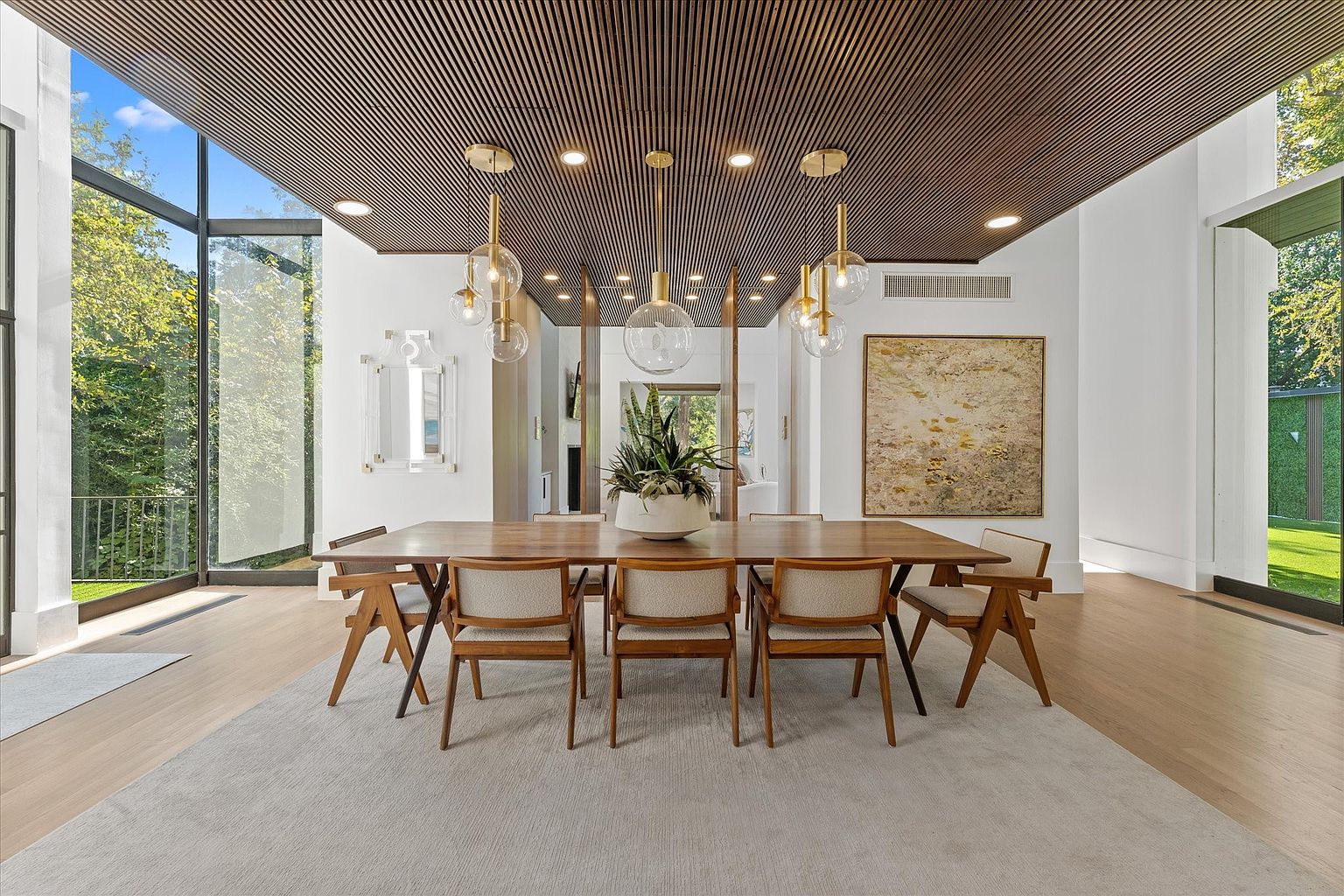 This is a dining room featuring a long wooden table with six chairs, set on a large neutral-toned rug. Above the table hang three modern pendant lights with glass globes. The room has large windows offering natural light and a view of the outdoors, complemented by a unique wooden slat ceiling and a piece of art on the wall.