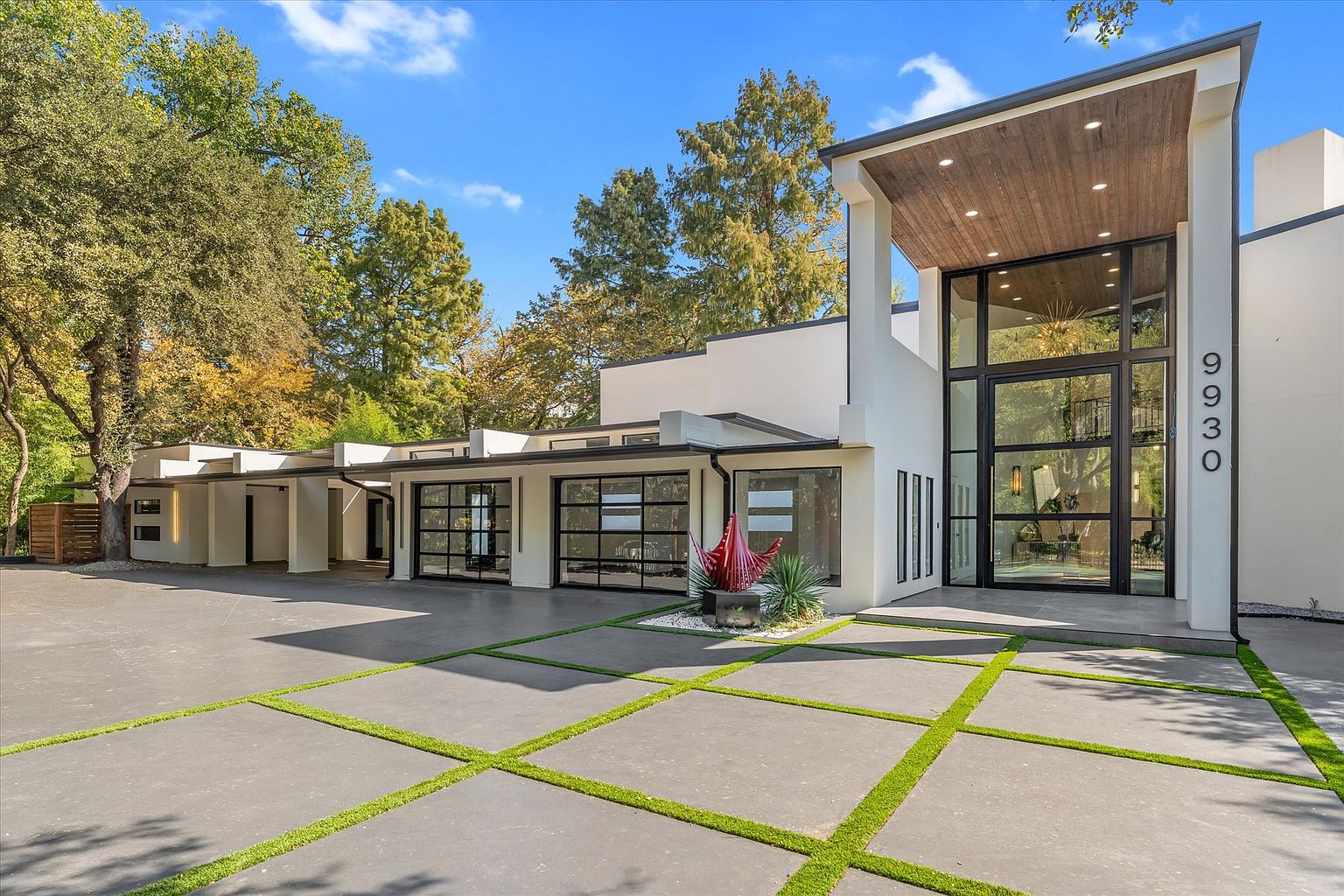 This is a striking front view of a modern home, showcasing its clean lines, flat roof, and expansive glass windows. The property features a spacious driveway with grass accents, leading to a multi-car garage and a grand entryway with a wooden-paneled overhang. The architectural style is contemporary, emphasizing natural light and a seamless blend of indoor and outdoor spaces.