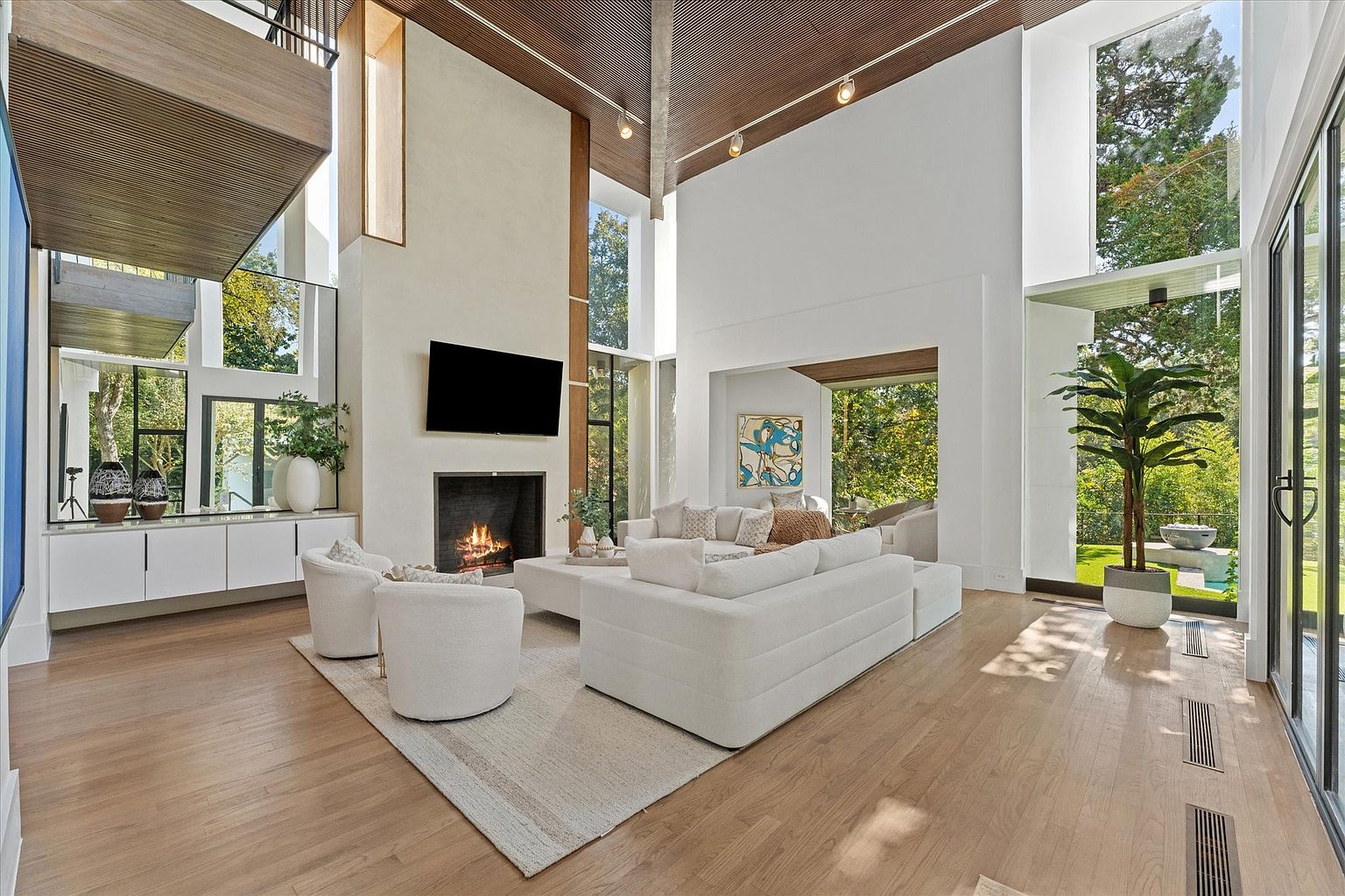 This is a spacious and modern living room featuring high ceilings, large windows, and a fireplace with a mounted TV above. The room is furnished with a white sectional sofa, two armchairs, and a light-colored area rug. The hardwood floors and natural light create a bright and inviting atmosphere, perfect for relaxation and entertaining.