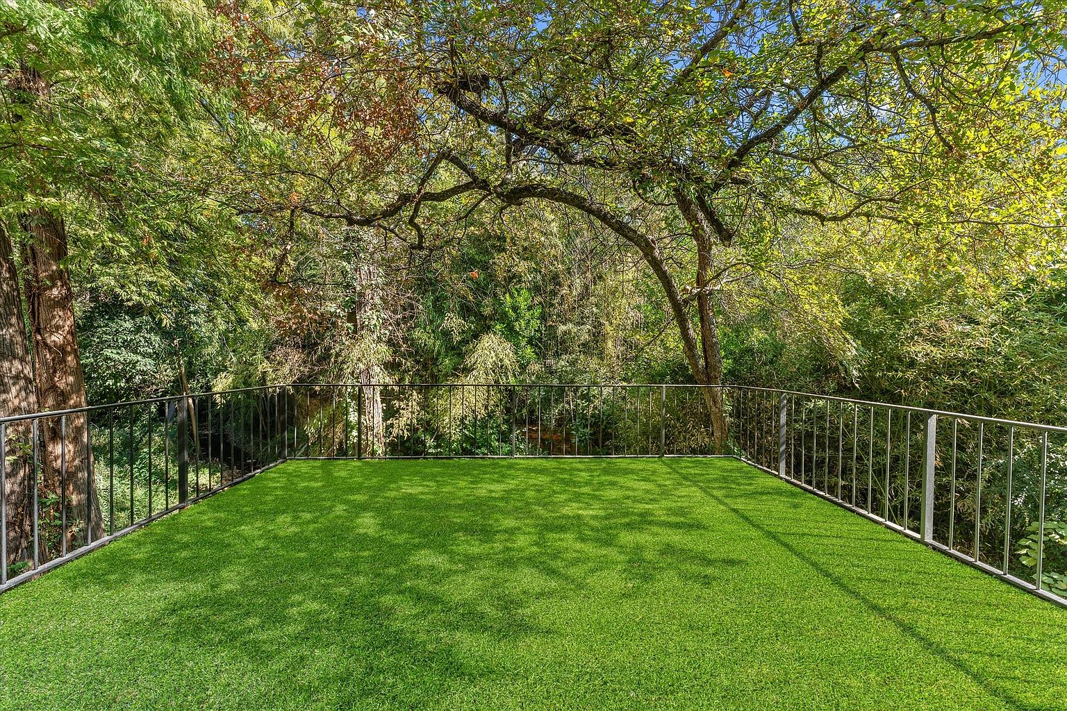 This image showcases a spacious outdoor deck or balcony area covered with artificial turf, offering a low-maintenance and visually appealing space. The deck is surrounded by a metal railing, providing safety and a clear view of the lush greenery and trees in the background. The scene evokes a sense of tranquility and privacy, making it an ideal outdoor retreat.