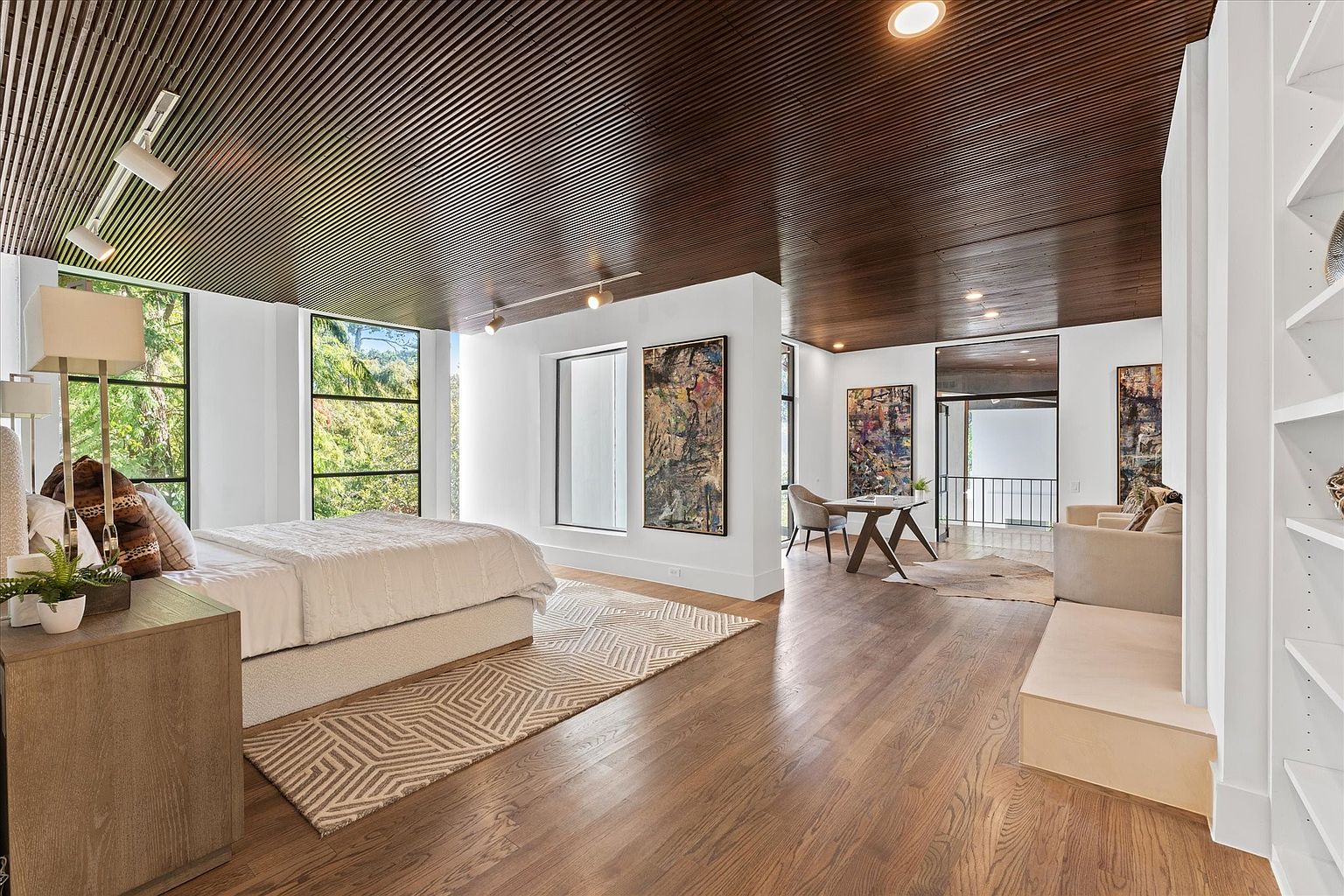 This is a spacious primary bedroom featuring a dark wood slat ceiling and light hardwood floors. The room is well-lit with natural light from large windows and features a bed, nightstand, rug, and artwork. The space also includes a seating area with a sofa and a desk, creating a luxurious and comfortable atmosphere.
