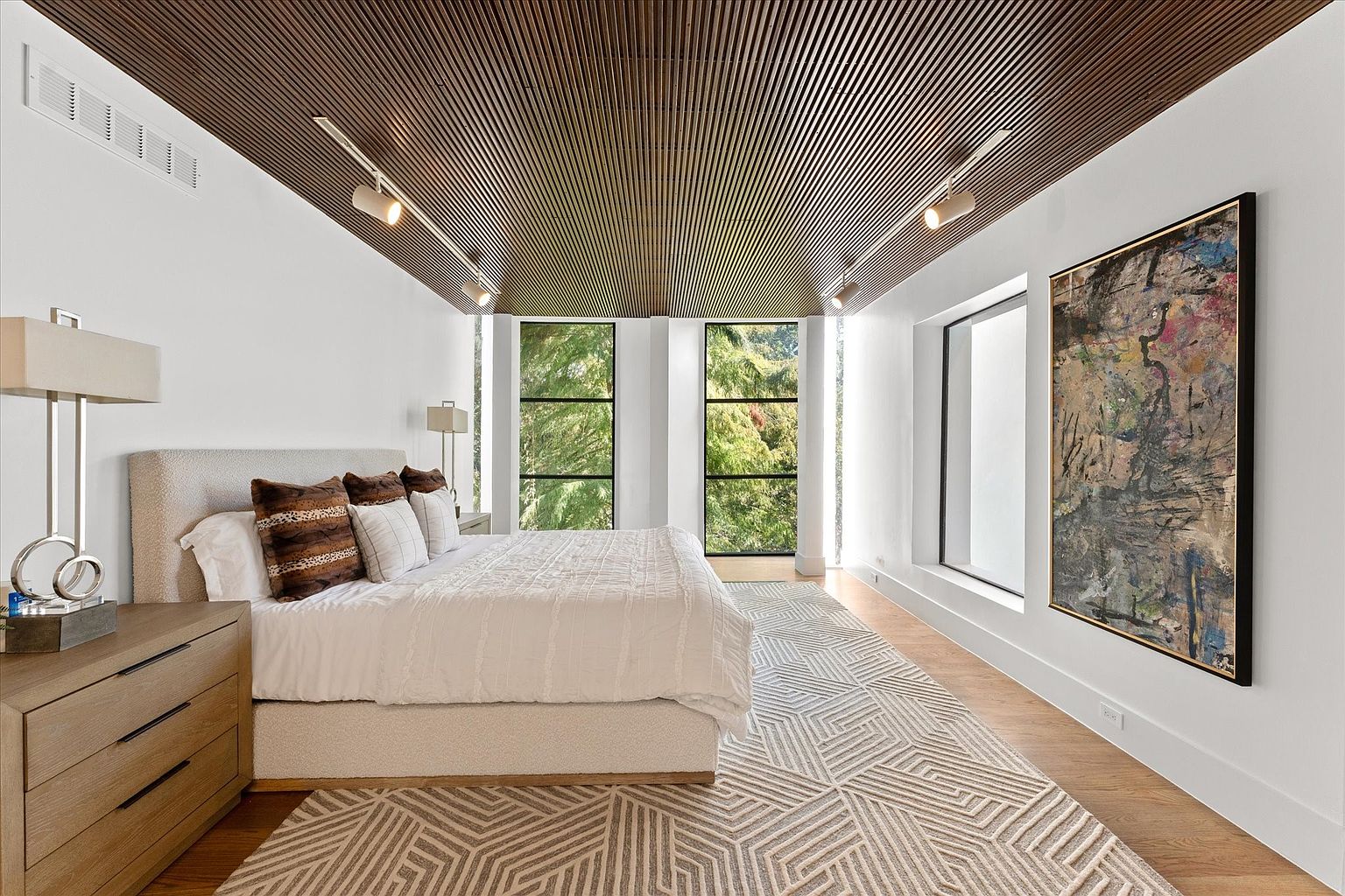 This is a primary bedroom featuring a king-size bed with a textured headboard, white bedding, and decorative pillows. The room has a modern design with a wood-paneled ceiling, track lighting, and large windows offering natural light and views of greenery. An abstract painting hangs on the wall, and a geometric patterned rug covers the hardwood floor.