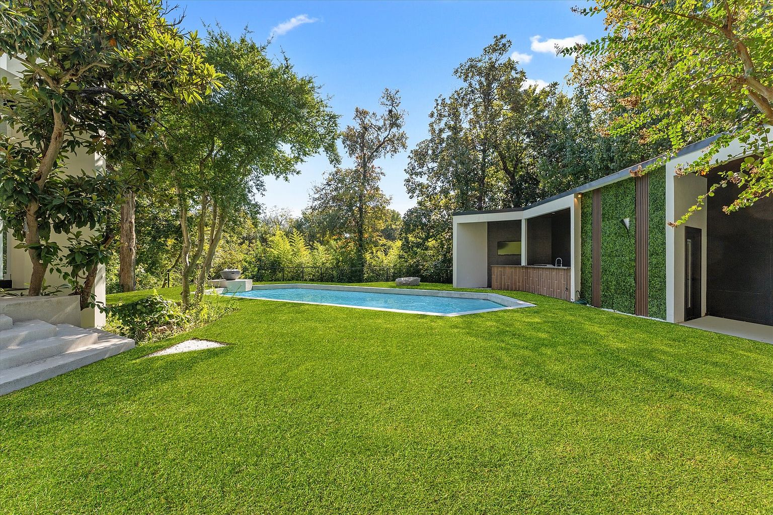 This image showcases a beautifully landscaped backyard featuring a well-maintained lawn, mature trees, and a modern pool. A contemporary pool house with a bar area and vertical garden adds a touch of luxury. The overall impression is one of tranquility and sophisticated outdoor living.