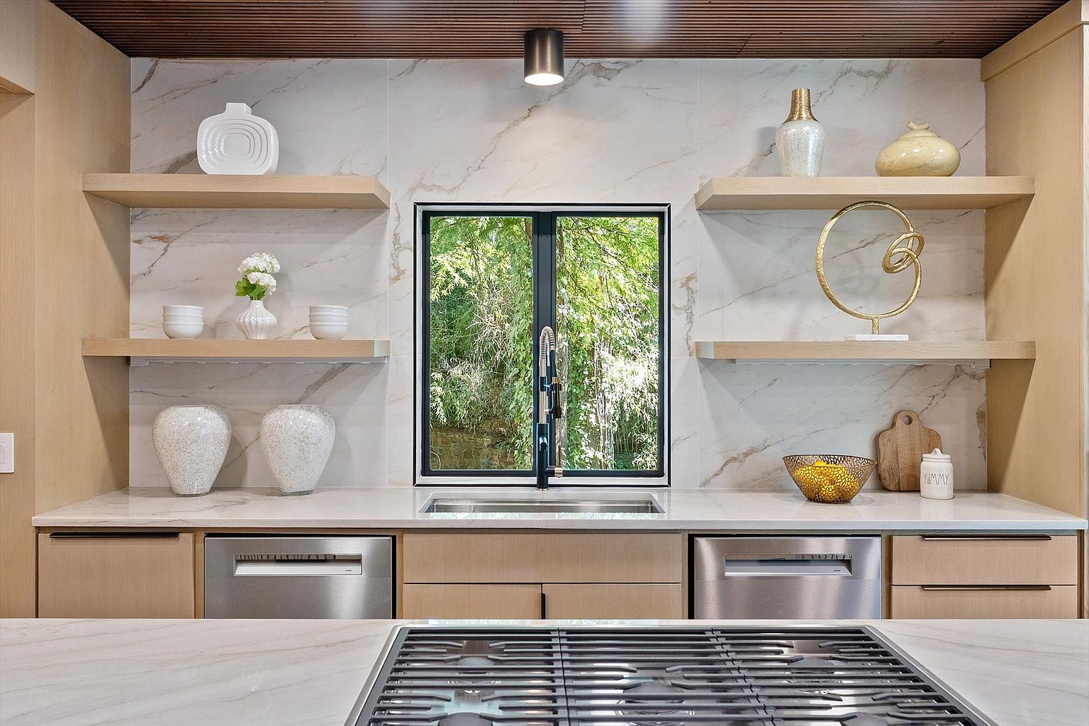 This is a well-lit kitchen featuring a window overlooking greenery, light wood cabinetry, and stainless steel appliances. The countertops are a light marble, and open shelving displays decorative items. The perspective is from the front, showcasing the stove and sink area.