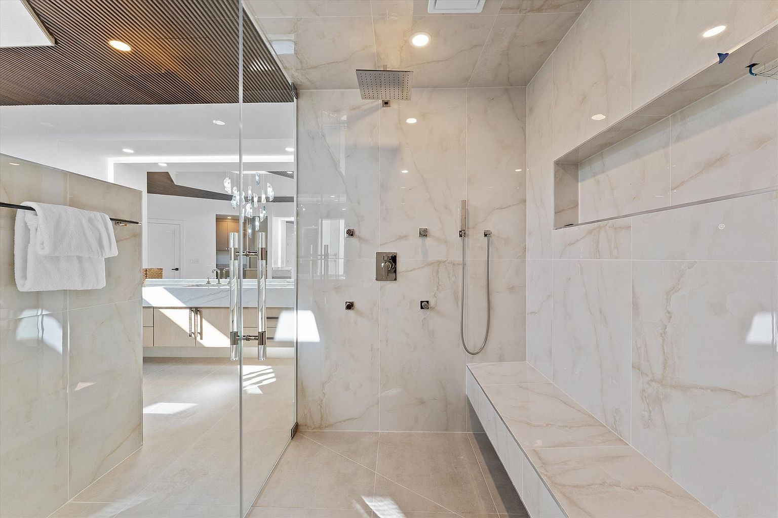 This is a luxurious primary bathroom featuring a spacious walk-in shower with marble-like tile, a rainfall showerhead, and a built-in bench. The shower is enclosed with glass, offering a view into the rest of the bathroom with a vanity and decorative lighting. The overall impression is clean, modern, and spa-like.