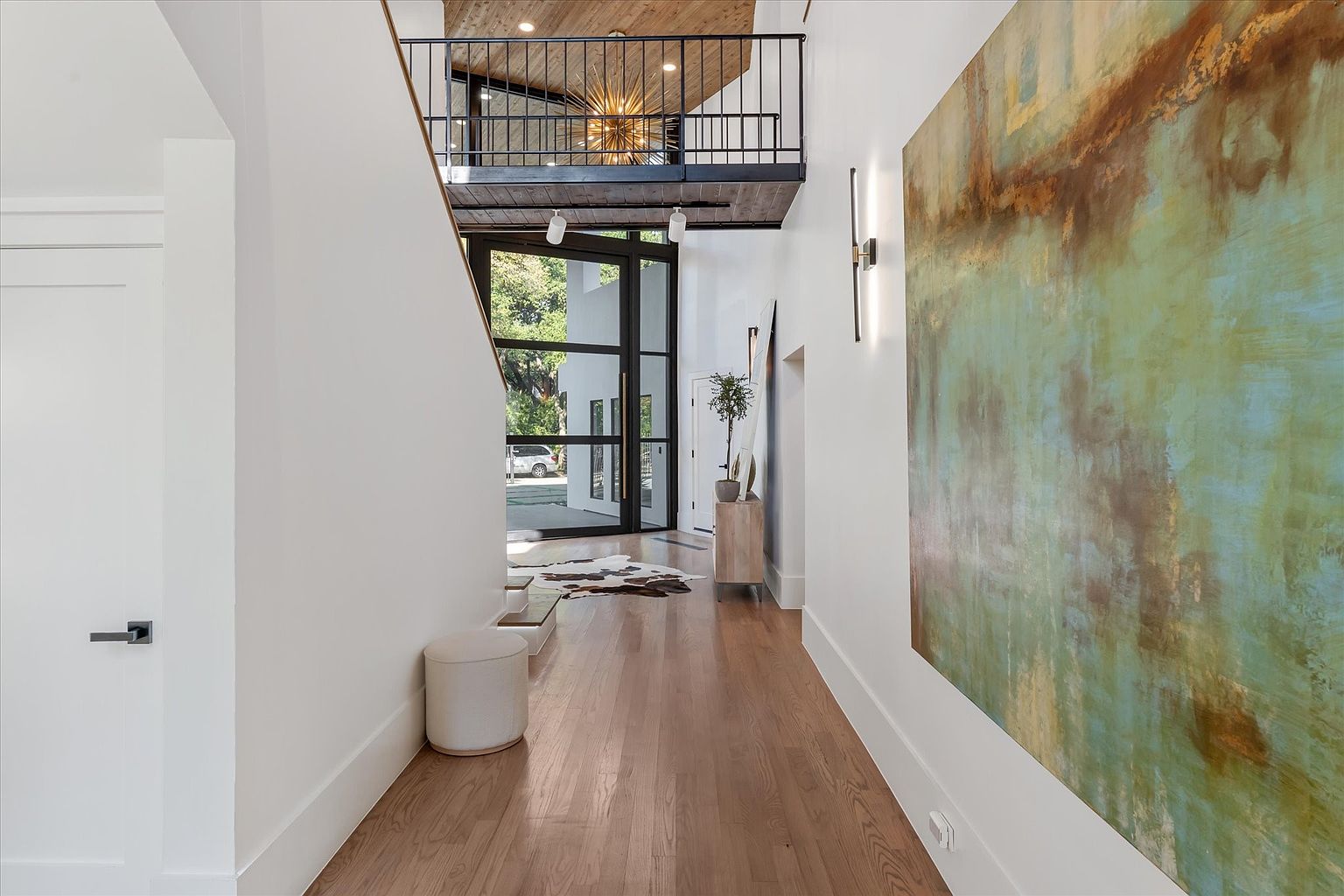 This interior shot showcases a modern hallway with hardwood floors and white walls, leading to a glass-paneled front door. A large abstract painting adorns the wall, complemented by a sleek sconce. A mezzanine overlooks the hallway, adding architectural interest and a sense of spaciousness.