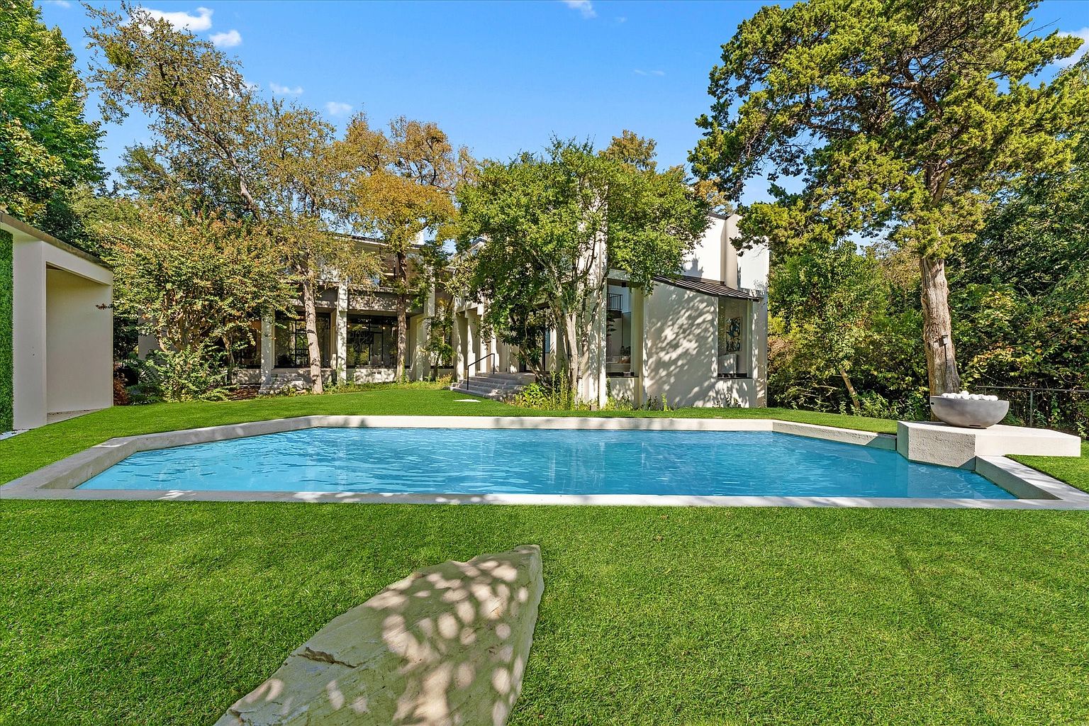 This image showcases a stunning backyard featuring a large, rectangular swimming pool with a modern design. The pool is surrounded by lush green grass and mature trees, creating a private and serene oasis. A contemporary home is visible in the background, complementing the clean lines of the pool area.
