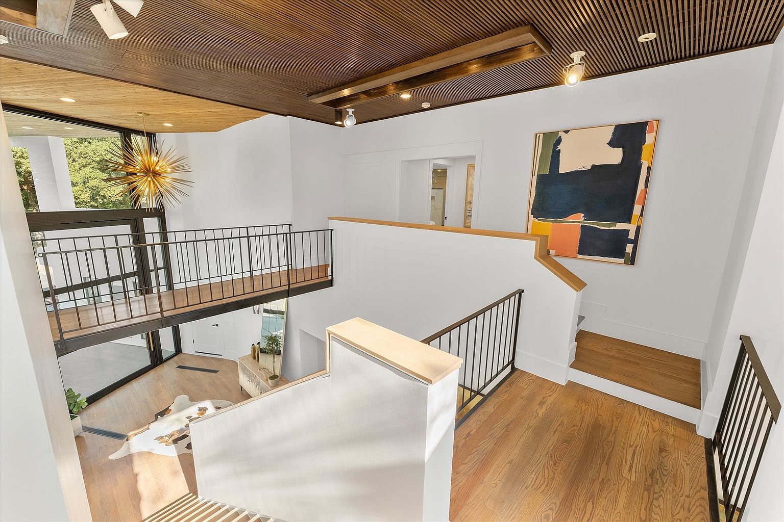 This interior shot showcases a modern hallway and staircase with a unique architectural design. The space features a wooden slat ceiling, white walls, and hardwood floors, complemented by black metal railings. A large abstract painting adds a touch of color, while a sputnik chandelier provides a focal point, creating a bright and airy atmosphere.