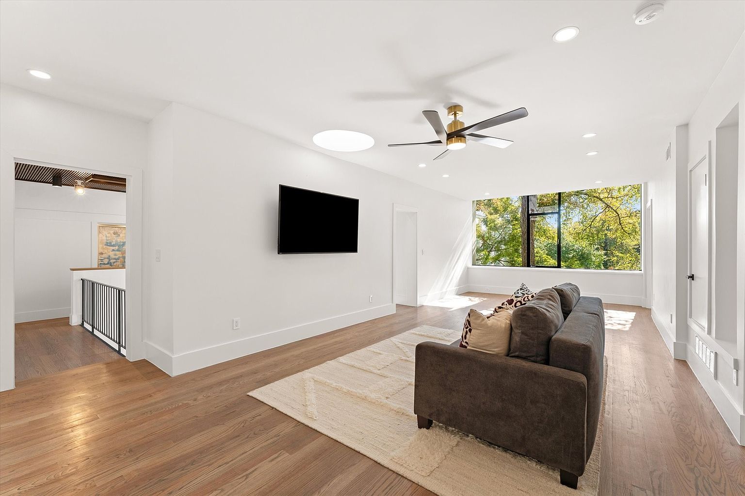 This is a bright and airy living room featuring hardwood floors, white walls, and a large window offering natural light. A gray sofa is positioned on a light-colored rug, facing a wall-mounted television. The room also includes a modern ceiling fan and recessed lighting, creating a comfortable and inviting atmosphere.