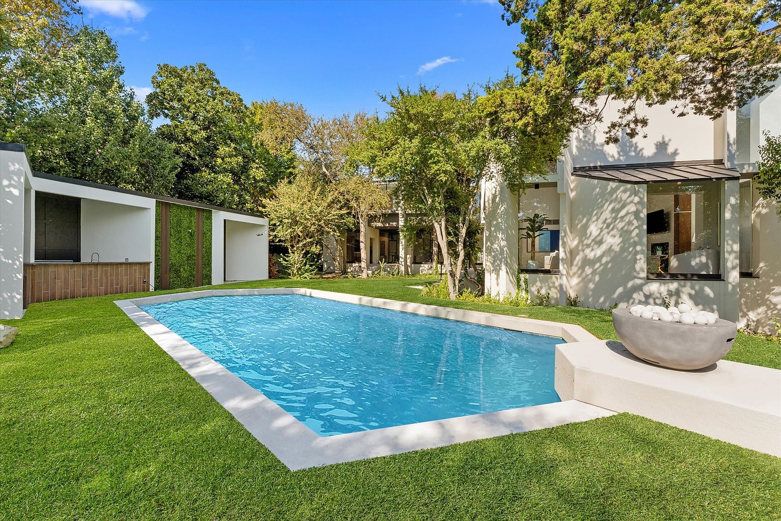 This image showcases a modern backyard with a rectangular pool as the focal point. The pool is surrounded by lush green grass and features a clean, white border. A contemporary outdoor structure with a wooden bar and vertical garden adds a stylish touch, while the main house is visible in the background, framed by mature trees. A modern fire pit sits near the pool.