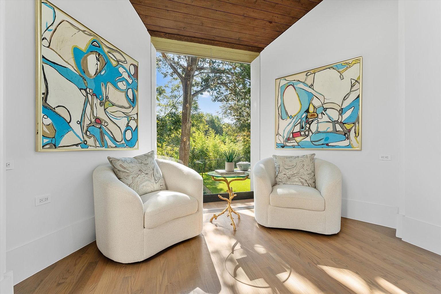 This interior shot showcases a bright and airy living room with two plush, cream-colored armchairs flanking a small, ornate side table. Large abstract paintings adorn the walls, adding a touch of modern art, while a large window frames a lush green outdoor view, creating a seamless connection with nature. The hardwood floors and natural light enhance the room's inviting and sophisticated ambiance.
