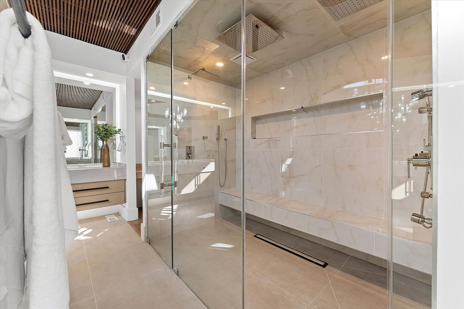 This is a luxurious primary bathroom featuring a spacious walk-in shower with glass enclosures, a built-in bench, and dual shower heads. The walls and floors are covered in light-colored marble tiles, creating a bright and elegant atmosphere. A modern vanity with sleek drawers and a vessel sink is visible in the background, complemented by a decorative plant.
