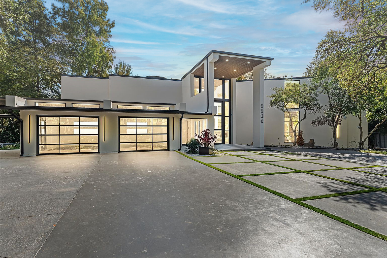 This image showcases the front exterior of a modern home. The architectural design includes a striking entryway with a high ceiling, complemented by a multi-car garage featuring glass doors. The property boasts a contemporary driveway design with integrated greenery, adding to the home's curb appeal.