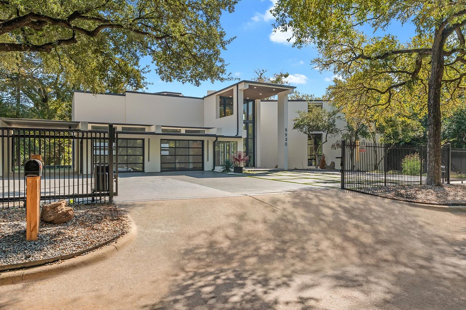This is a front exterior view of a modern, two-story home with a gated entrance. The house features a white facade with black accents, a flat roof, and large glass garage doors. The property is surrounded by mature trees, and the driveway leads to the front of the house, creating a welcoming and luxurious impression.