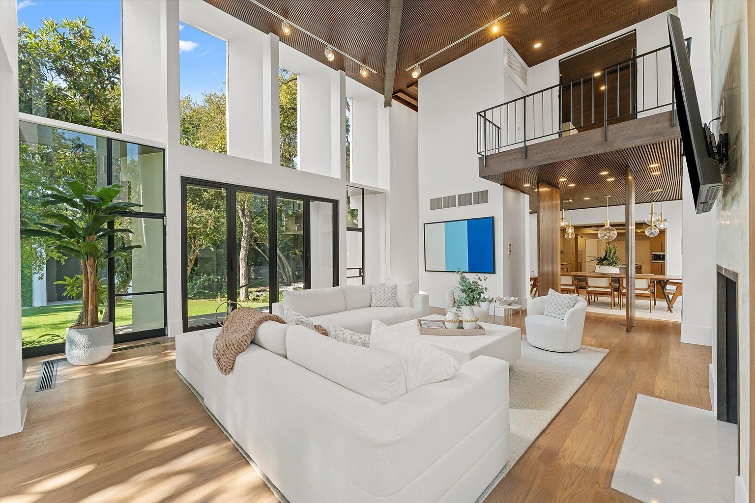 This is a bright and airy living room featuring a soaring ceiling, large windows, and a modern aesthetic. The room is furnished with a large white sectional sofa, a white area rug, and a contemporary coffee table. The open floor plan seamlessly connects the living room to the dining area, creating a spacious and inviting atmosphere.