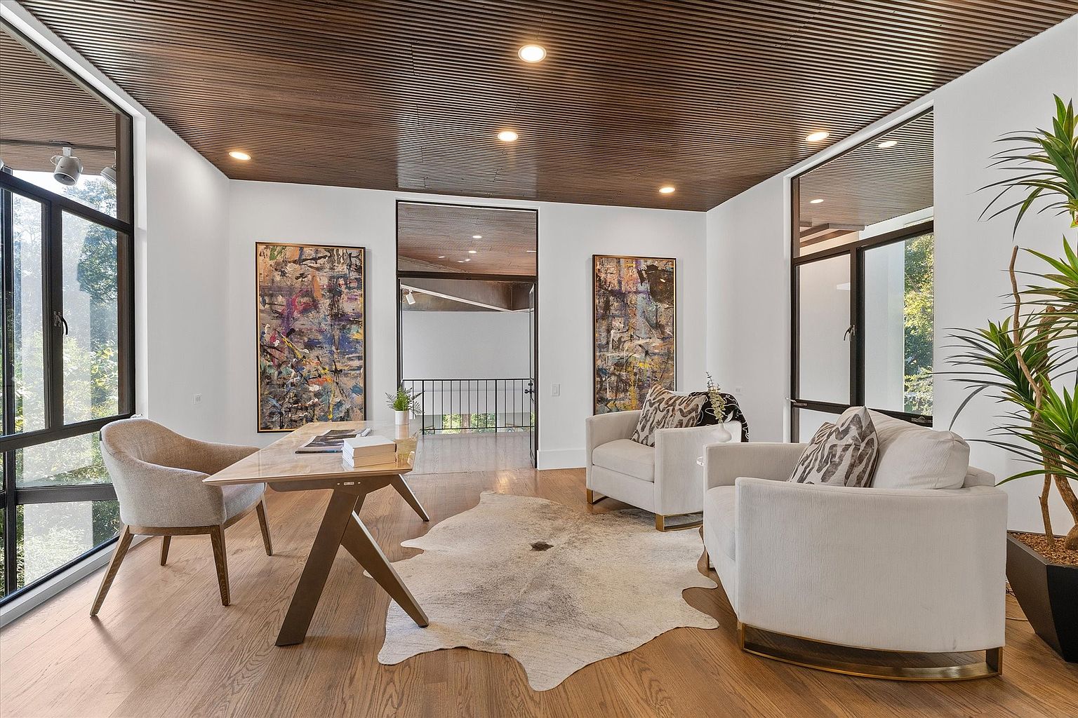 This is an interior shot of a modern office or study. The room features a wooden slat ceiling, hardwood floors, and white walls. There is a desk with a chair, two armchairs, and abstract art on the walls. Large windows provide natural light, and a cowhide rug adds texture to the space.