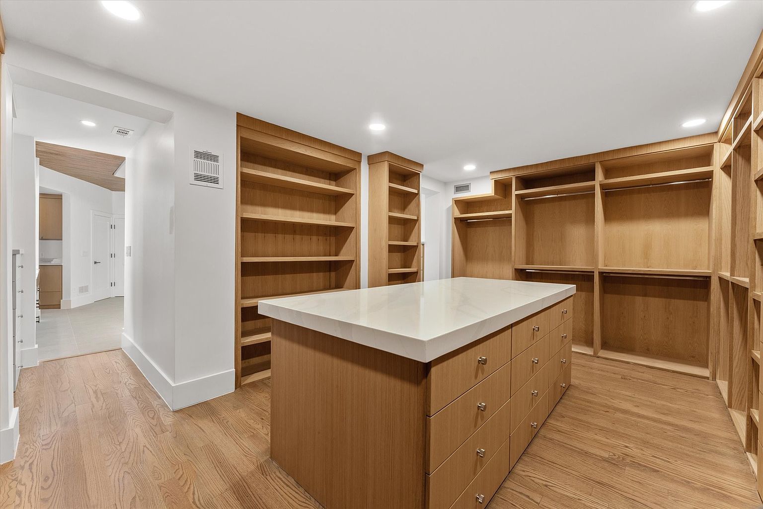 This is a spacious walk-in closet featuring custom wood shelving and drawers. A large island with a light countertop sits in the center, providing additional storage and workspace. The hardwood flooring and ample lighting create a luxurious and organized space.