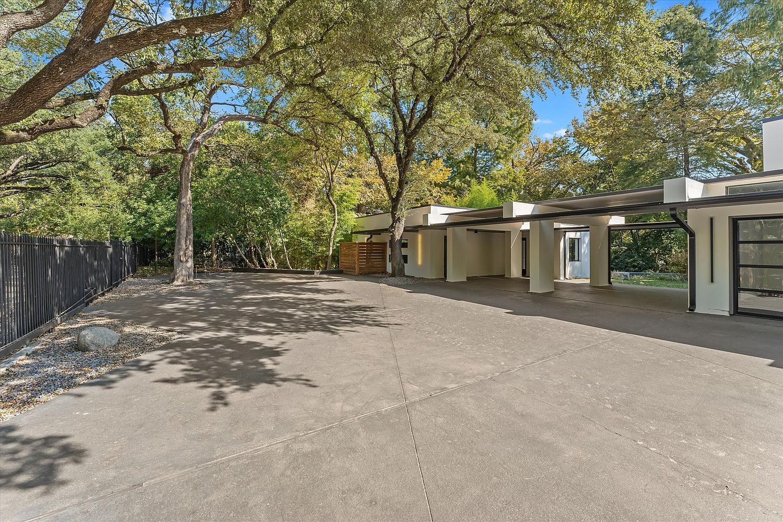 This image showcases the front exterior of a modern home, emphasizing its sleek architectural design with a covered carport supported by white pillars. The expansive concrete driveway provides ample parking space, while mature trees offer shade and enhance the property's curb appeal. A black metal fence adds a touch of security and defines the property line.