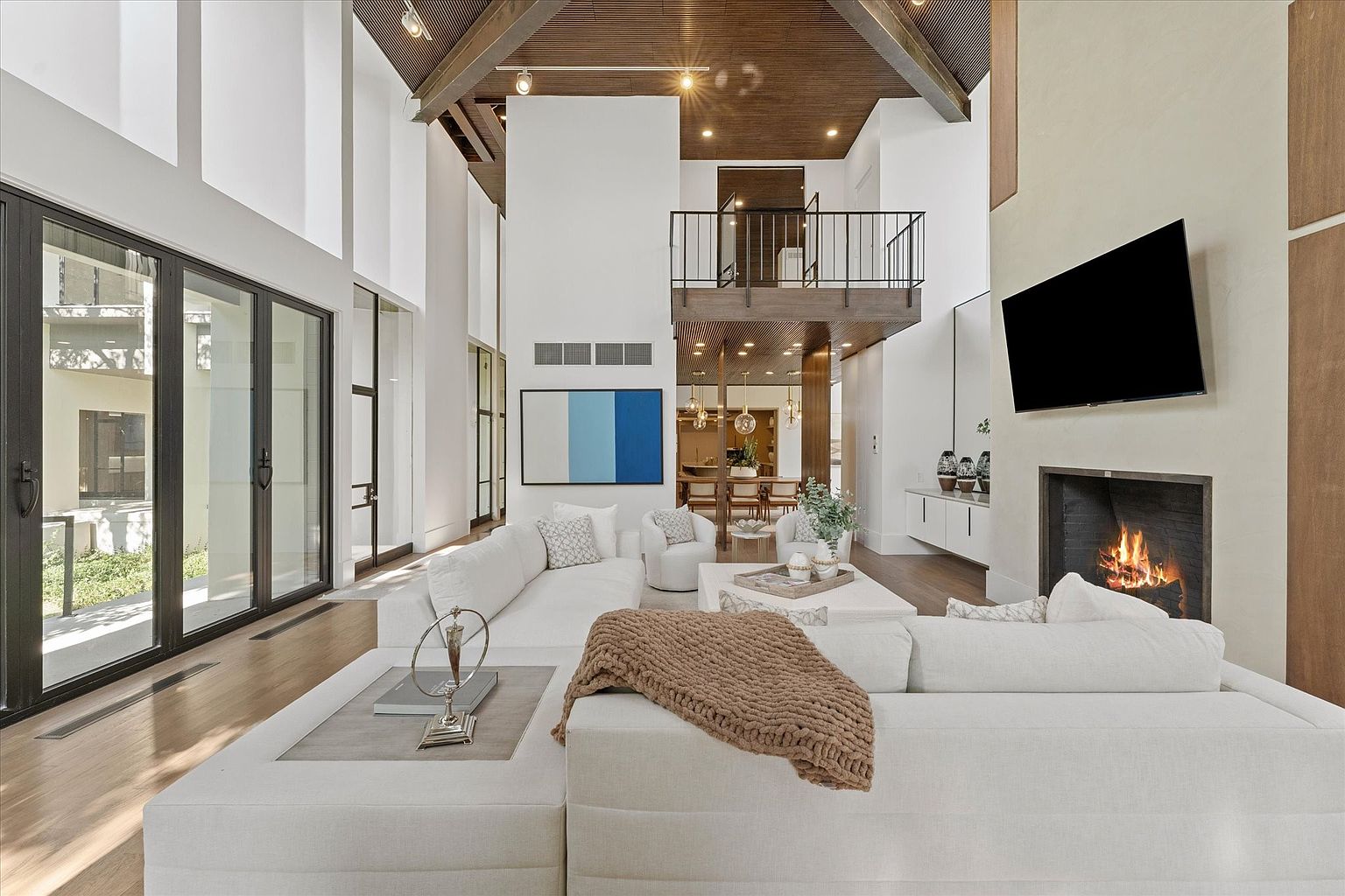 This is a spacious, modern living room with high ceilings and a striking architectural design. The room features a large white sectional sofa, a fireplace with a mounted television, and expansive windows that provide ample natural light. A second-story balcony overlooks the living area, adding to the grandeur of the space.