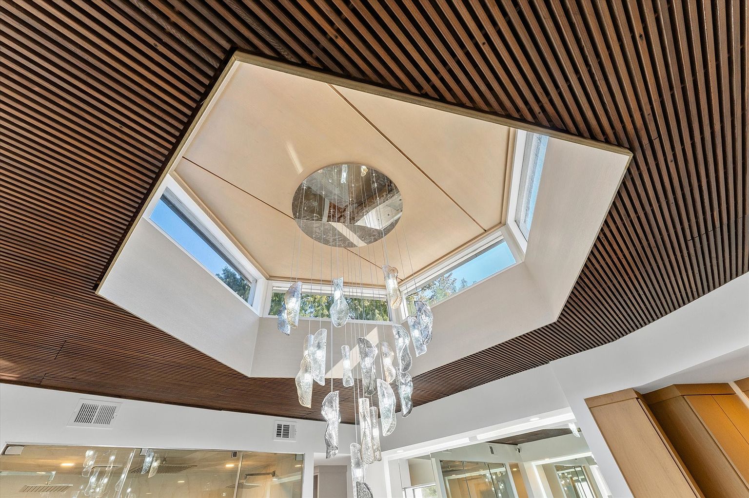This image showcases a unique architectural detail: a modern chandelier hanging beneath a geometric ceiling feature with integrated windows. The ceiling is a combination of wood slats and a light-colored, possibly fabric-covered, recessed area. The chandelier itself is composed of irregularly shaped glass elements, creating a focal point that blends contemporary design with natural light.