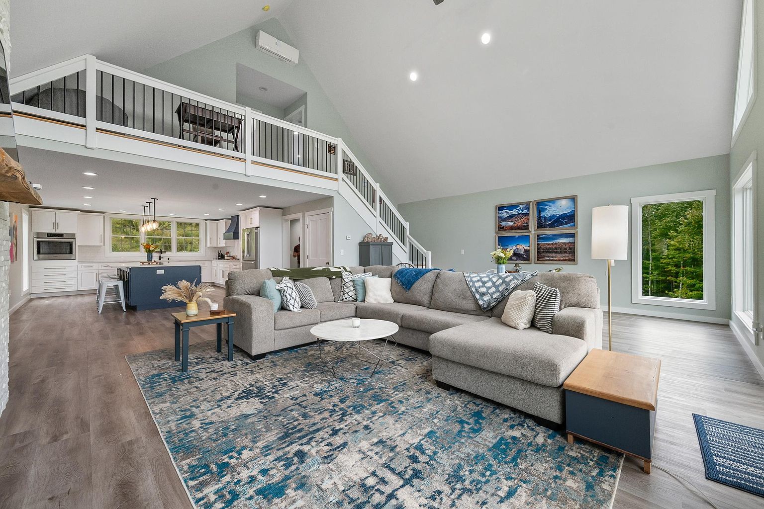 This spacious, open-concept living area features a vaulted ceiling and a neutral color palette, seamlessly connecting to a modern kitchen with a large island. A plush, L-shaped sectional sofa sits atop a textured blue and grey area rug, creating a comfortable focal point for the room. The space is brightened by natural light from large windows and includes a lofted walkway overlooking the main living area, enhancing the airy and expansive feel of the home.