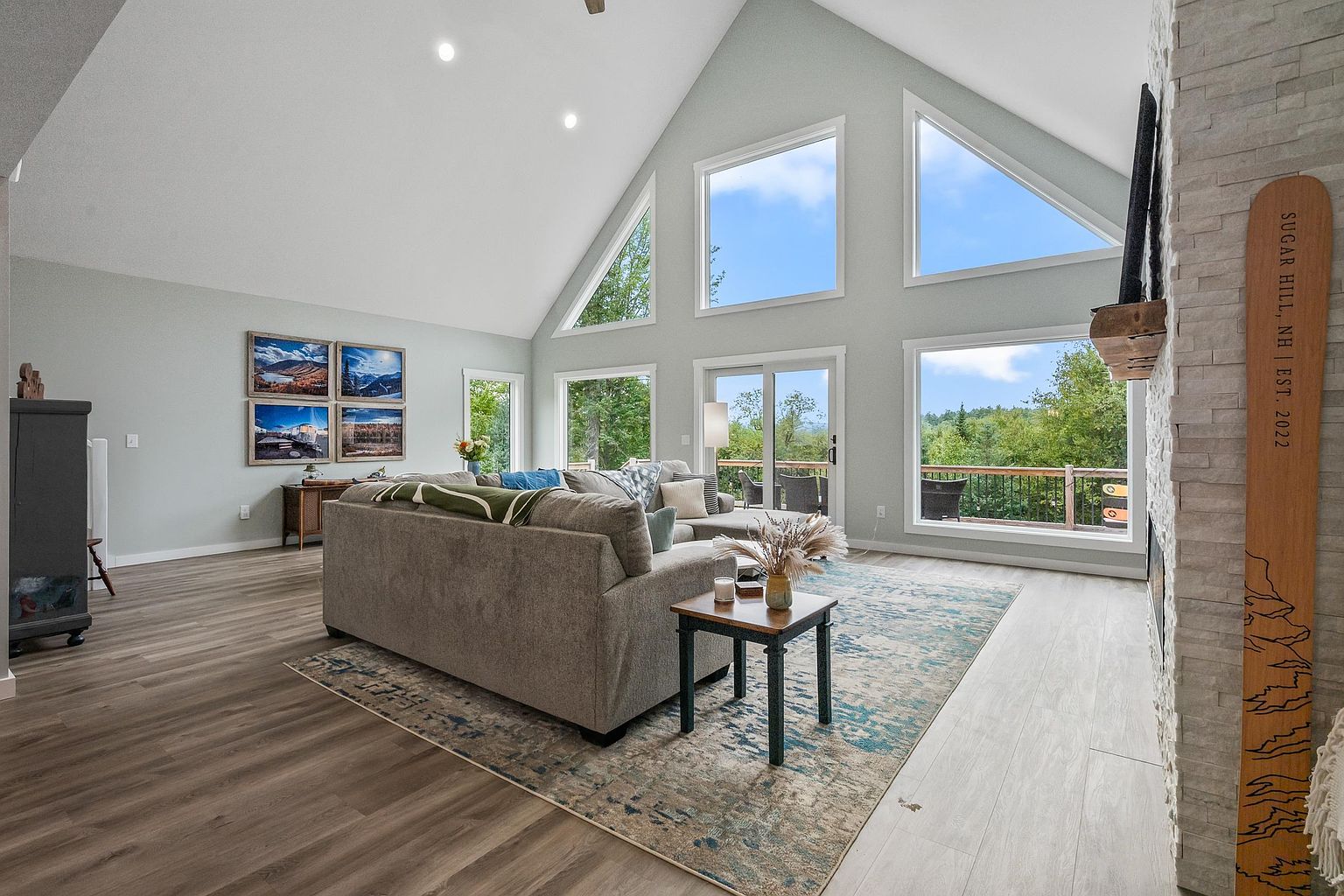 This spacious living room features a vaulted ceiling and a large wall of windows that provide abundant natural light and views of the surrounding landscape. A comfortable gray sectional sofa is centered on a patterned area rug, complemented by a small side table and a decorative wooden ski mounted on the stone-clad chimney breast. The open-concept layout creates an airy, inviting atmosphere, perfect for relaxing while enjoying the scenic outdoor vistas.