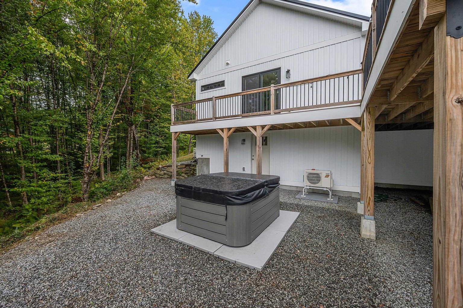This rear view of a modern white-sided home features a spacious elevated deck with black metal railings supported by wooden posts. Below the deck, a grey hot tub with a cover sits on a concrete pad, surrounded by a gravel yard that transitions into a lush, wooded forest. The scene captures a peaceful, private outdoor living space ideal for relaxation.