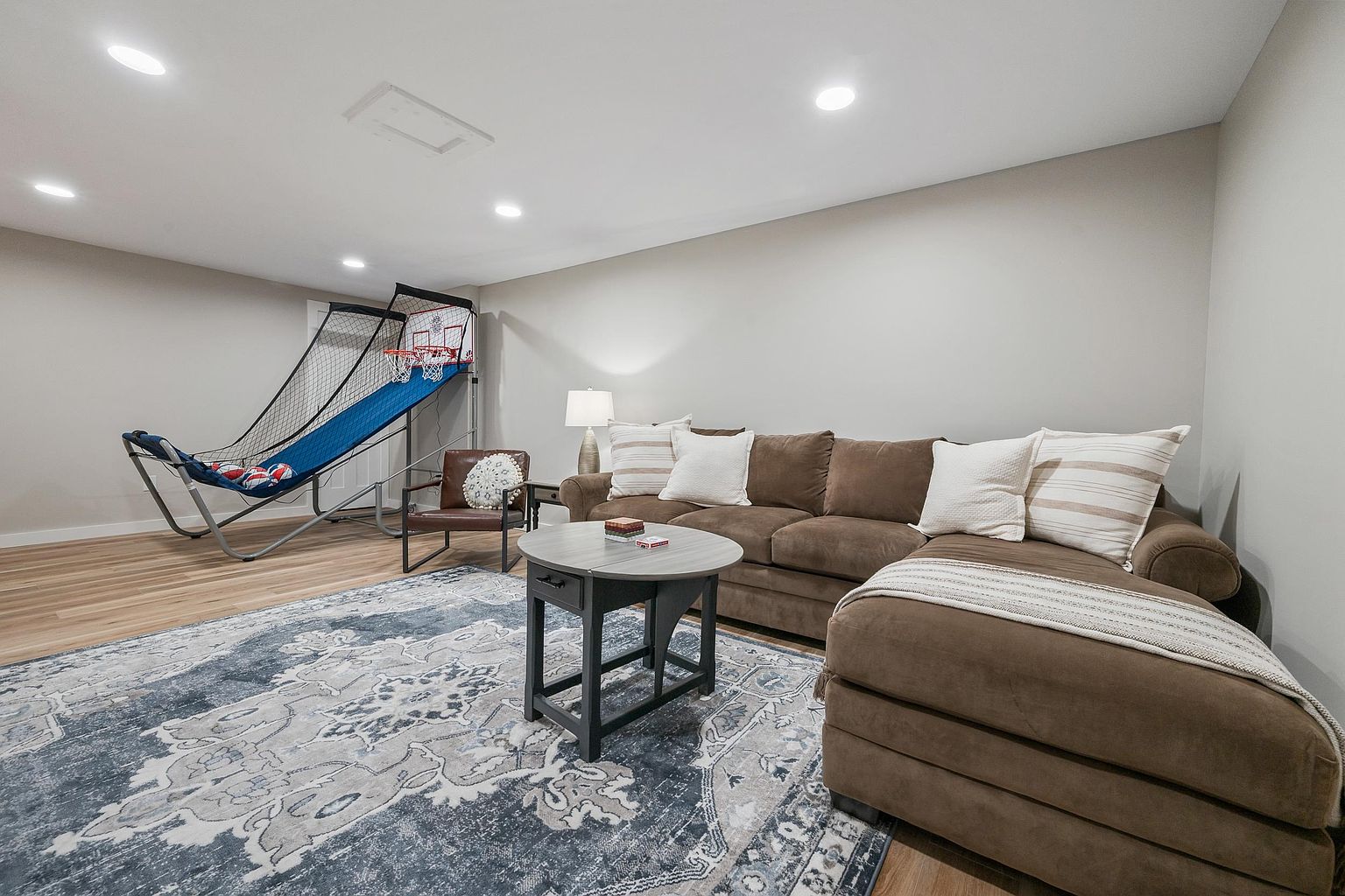 This finished basement space features light wood-look flooring and a large, comfortable brown sectional sofa adorned with neutral-toned throw pillows. A circular coffee table sits atop a prominent blue and grey patterned area rug, while a basketball arcade game is positioned in the corner, adding a recreational element to the room. The space is illuminated by recessed lighting, creating a bright and inviting atmosphere suitable for a family lounge or game room.